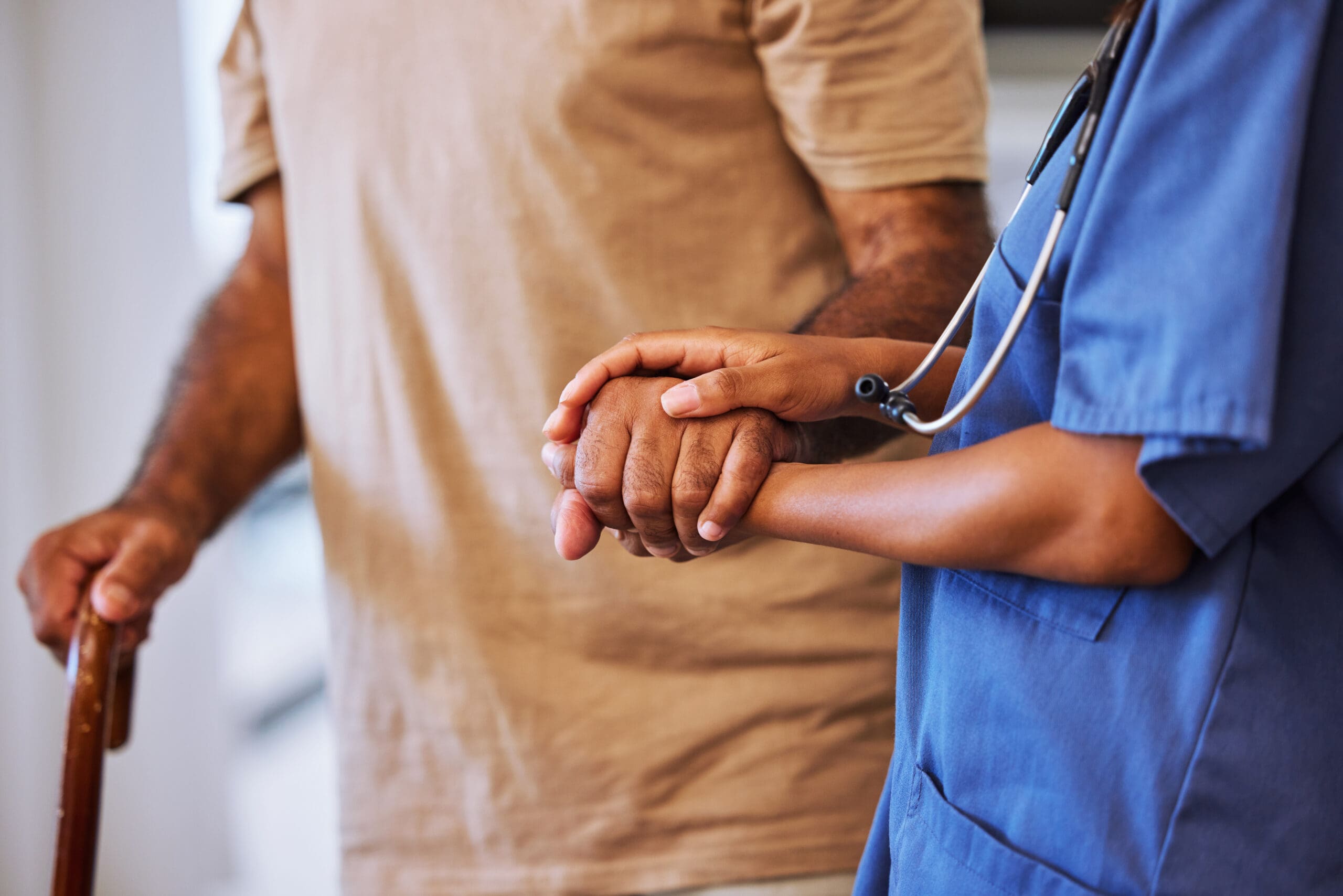 A healthcare professional in blue scrubs gently holding the hand of an elderly patient, who is using a cane. The patient's arm is partially visible, showing age spots and a beige shirt.