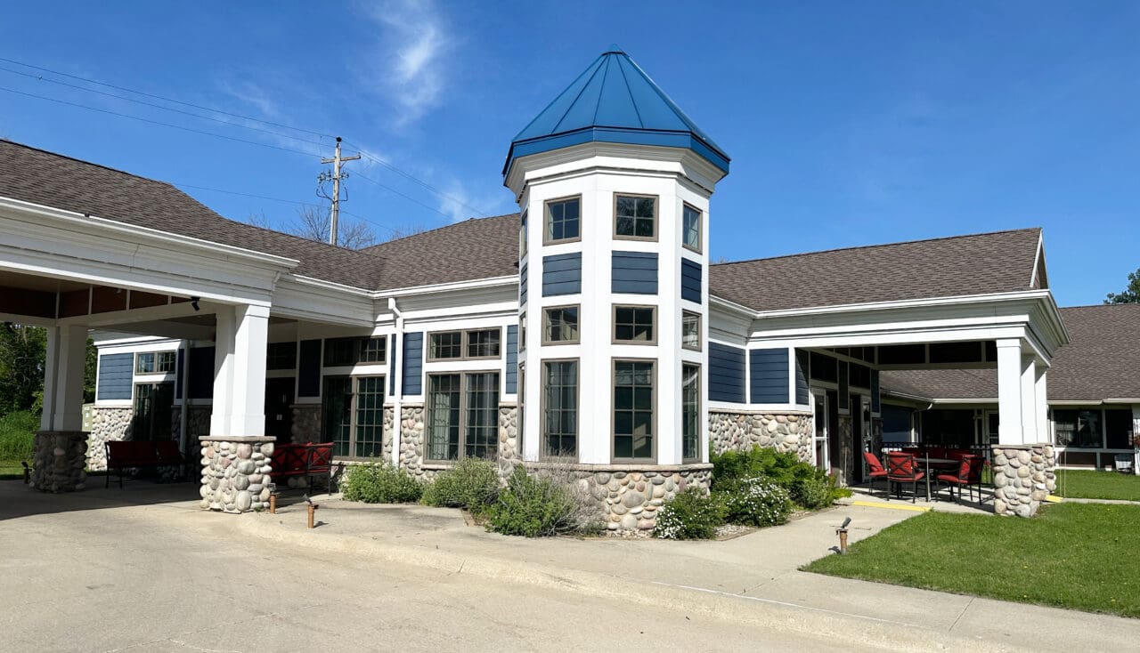 A single-story building with a central tower featuring multiple windows, blue siding with white trim, stone accents at the base, and a brown shingle roof. There are outdoor seating areas with red chairs and green landscaping in front.