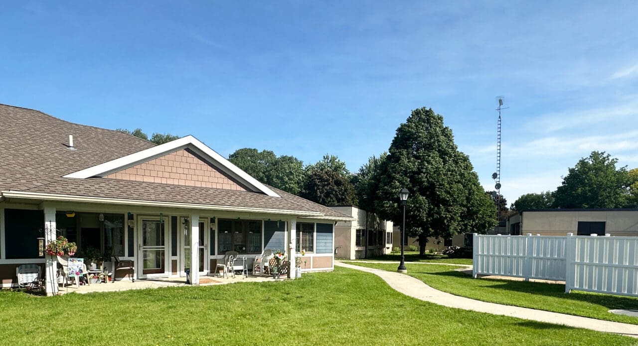 A single-story building with a brown roof, screened porch, and blue siding, surrounded by a well-maintained grassy lawn, a curved concrete walkway, a white fence, a large tree, and a clear blue sky.