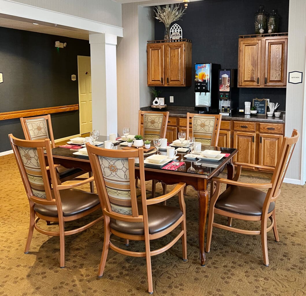 A dining area with a dark wood table set for a meal, surrounded by six cushioned wooden chairs with patterned fabric. In the background, there is a kitchen area with wooden cabinets, a juice dispenser, and a coffee machine. The table is set with plates, cups, glasses, and utensils.