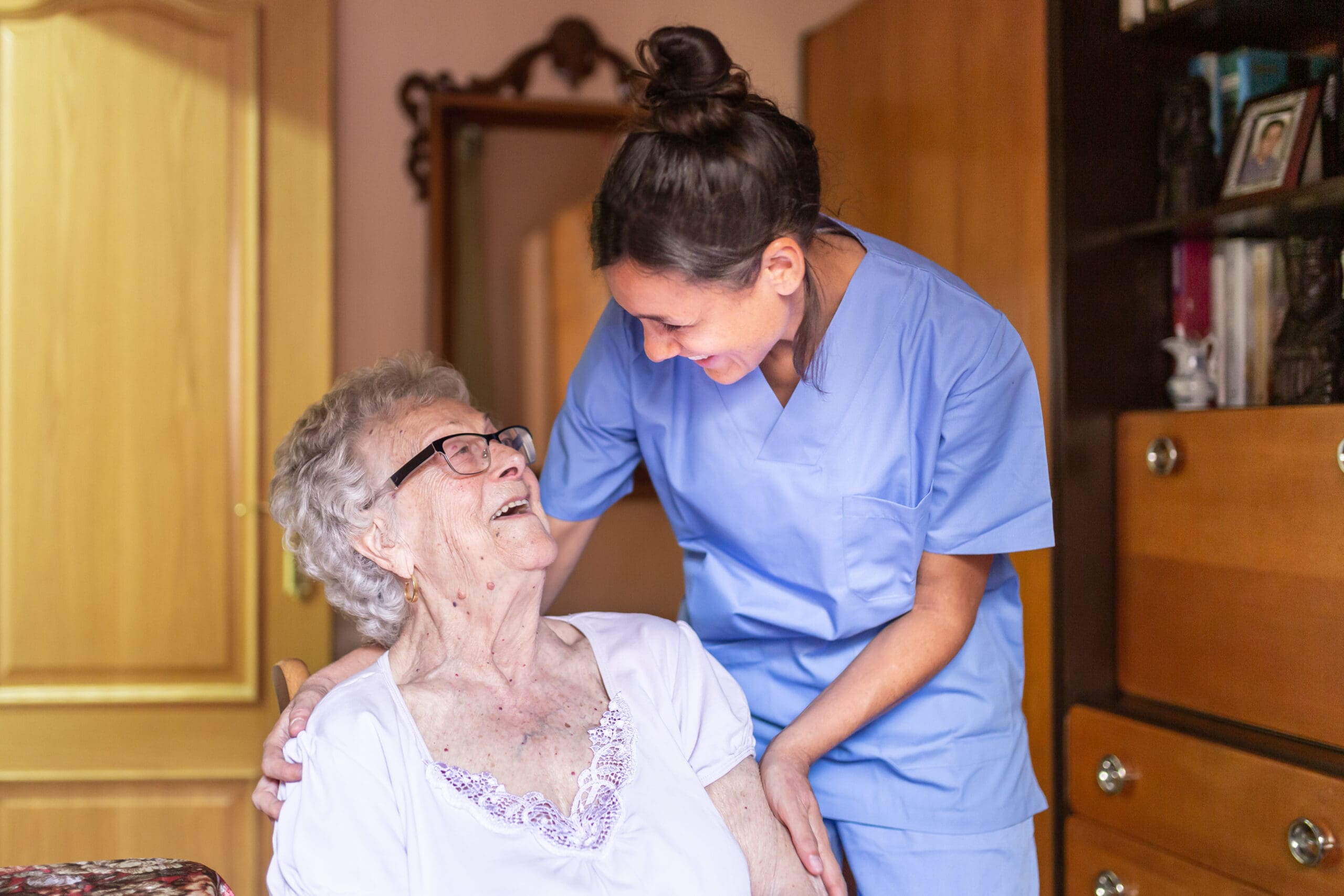 A healthcare worker in blue scrubs is smiling and leaning over an elderly woman with gray hair, glasses, and a white top, who is sitting and looking up at her with a joyful expression. They are sharing a warm, happy moment indoors.
