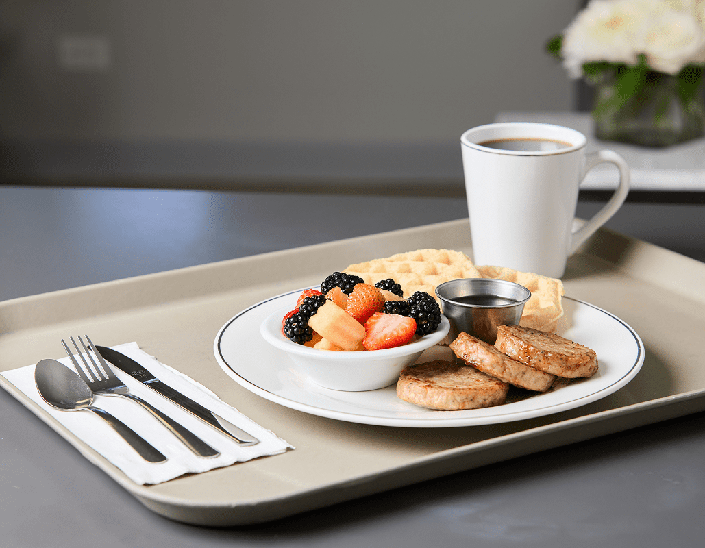 A tray with a breakfast meal includes a white bowl of mixed berries and melon, a plate with sausage patties, a waffle, and a small container of syrup, along with a cup of coffee.
